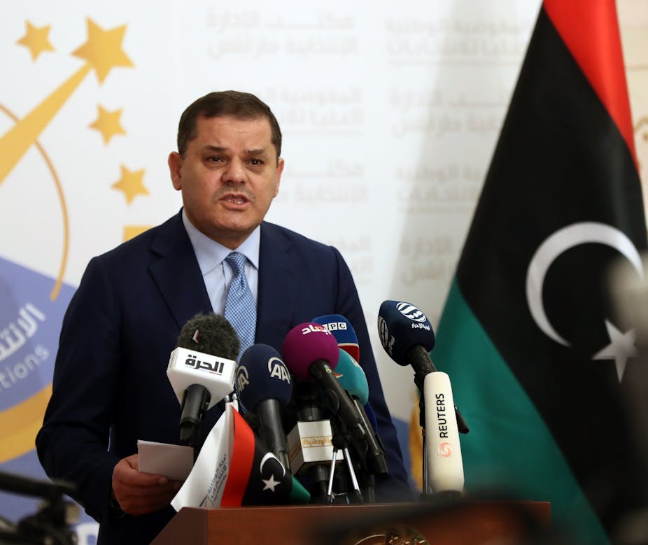 A man wearing a suit and tie speaks in front of several microphones. The Libyan flag is on his left.