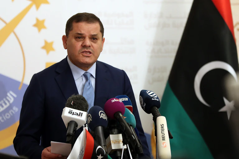 A man wearing a suit and tie speaks in front of several microphones. The Libyan flag is on his left.