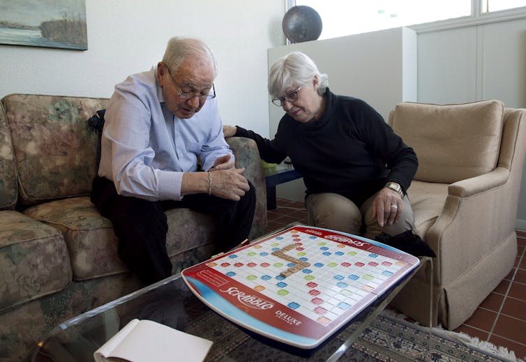 A man with grey hair and a woman with grey hair look down at a Scrabble board on a table in front of them.