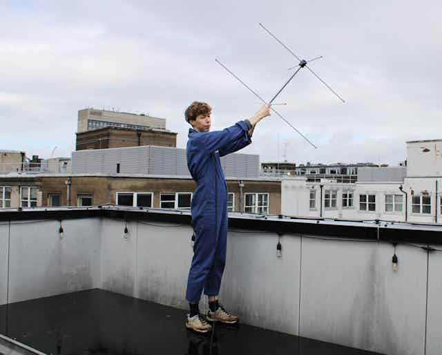 A person in blue overalls holds an antenna aloft on a roof in London.