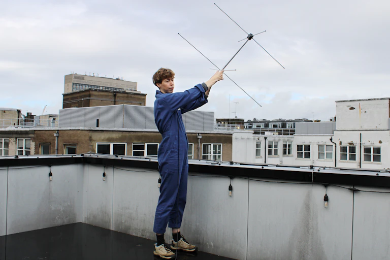 A person in blue overalls holds an antenna aloft on a roof in London.