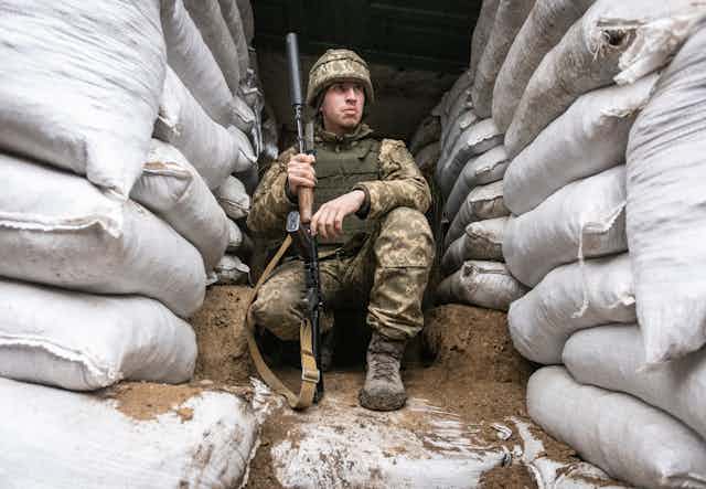 A soldier in combat gear and carrying a weapon in a military trench surrounded by sandbags.