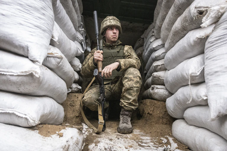 A soldier in combat gear and carrying a weapon in a military trench surrounded by sandbags.