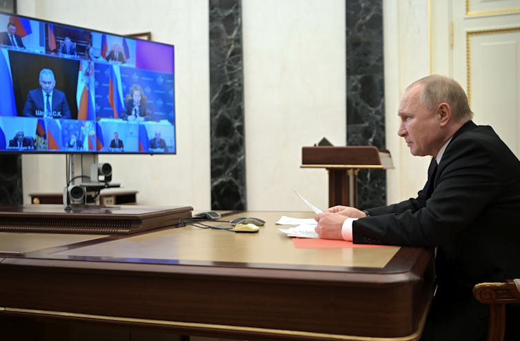 A bald man sits at a desk looking at a large computer monitor.
