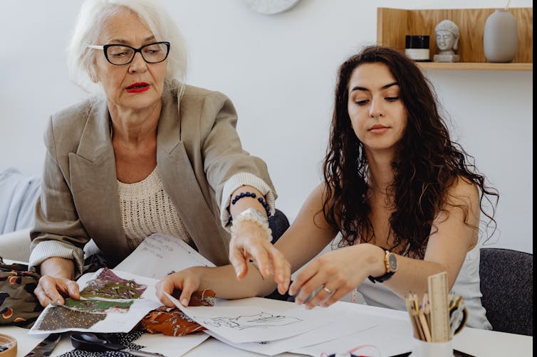 μια ηλικιωμένη γυναίκα με γκρίζα μαλλιά και γυαλιά δείχνει έγγραφα μπροστά σε μια νεότερη, μελαχρινή γυναίκα