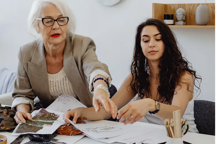 an older woman with grey hair and glasses points to documents in front of a younger, dark-haired woman