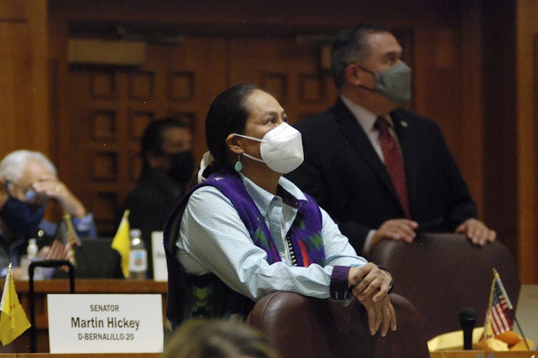 A woman with dark hair and wearing a mask and a colorful vest, stands behind a chair in a legislative meeting room.