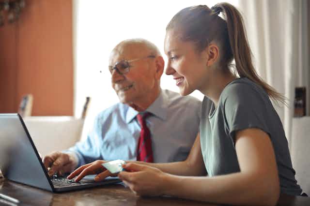 A young woman sits next to an elderly man in front of a laptop.