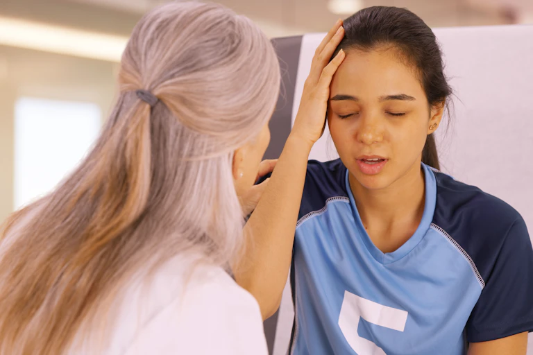 A teen girl in a sports uniform with her hand to her head, and a doctor seen from behind