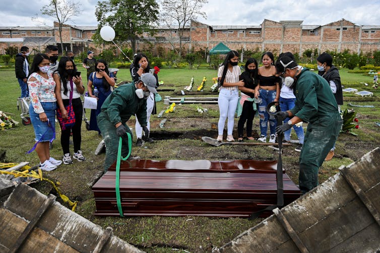 Two groups of young women stand together, looking over men securing the wooden coffins that they will lower into the ground.