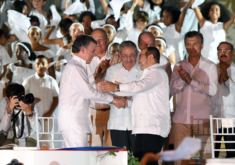 Colombian President Juan Manuel Santos shakes hands with former FARC leader Timoleon Jimenez. Both are dressed in white, and stand in front of a crowd of people also dressed in white