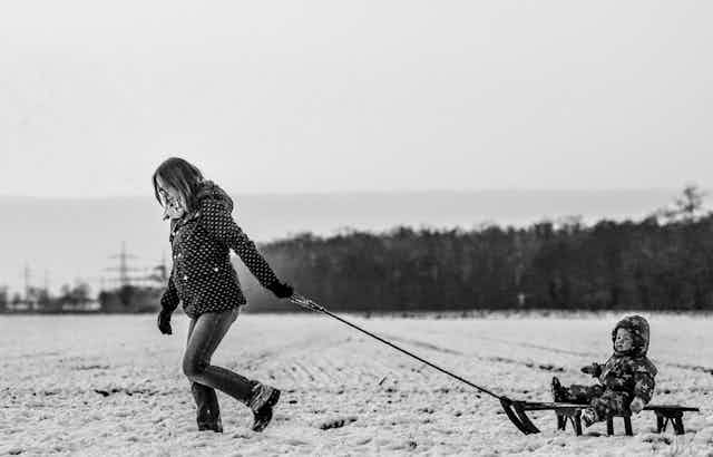 Black and white photo of a woman pulling a sled with a small child in a snowsuit through a snowy landscape