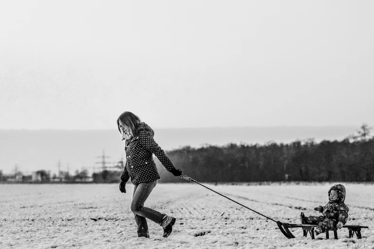Black and white photo of a woman pulling a sled with a small child in a snowsuit through a snowy landscape