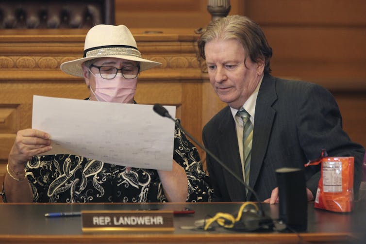 A woman in a straw hat and wearing glasses and a mask confers with a man about something on a piece of paper that the woman is holding.