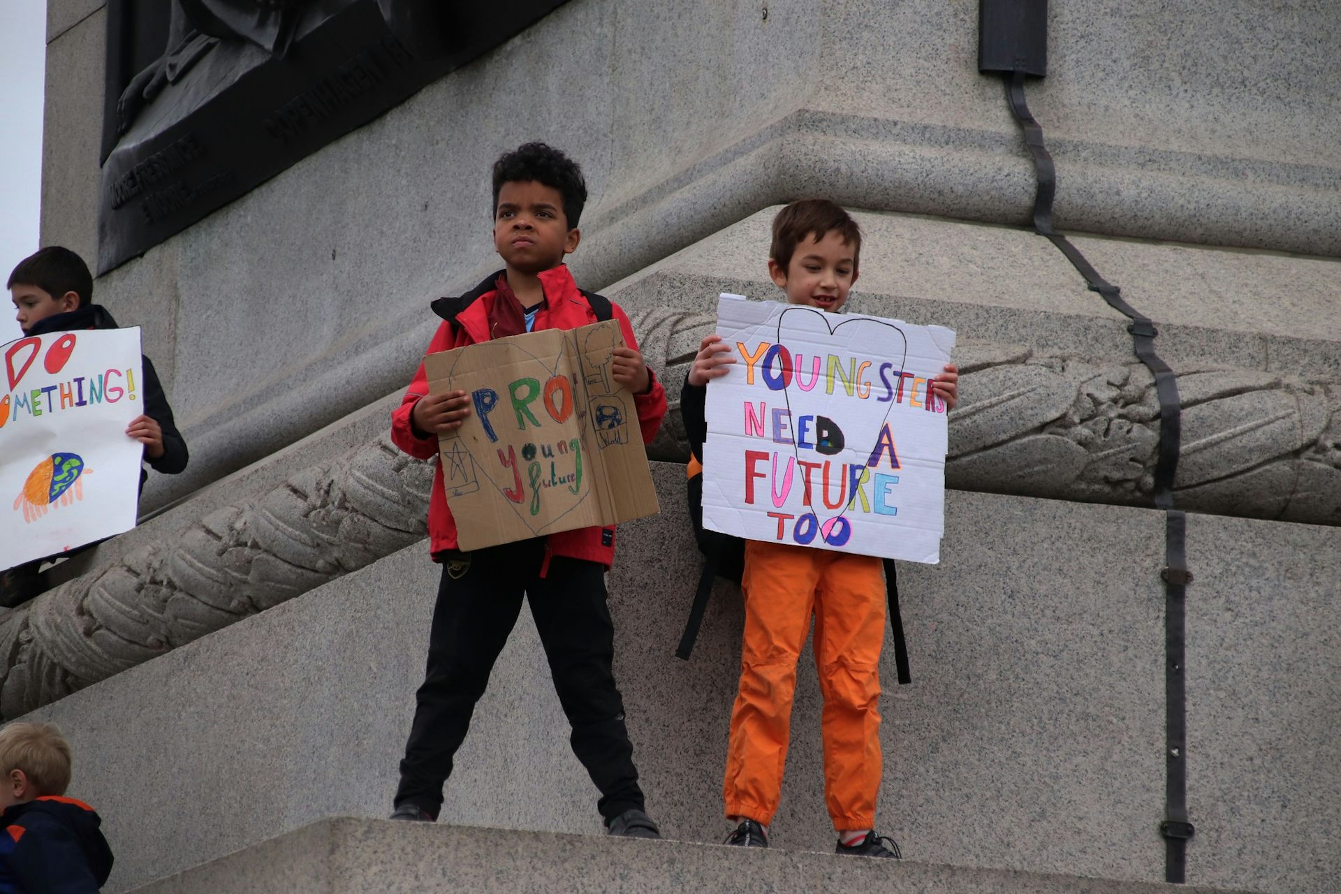 Children stand with protests signs on a statue; one sign says 'youngsters need a future too.'