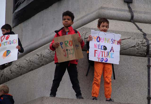 Children stand with protests signs on a statue; one sign says 'youngsters need a future too.'