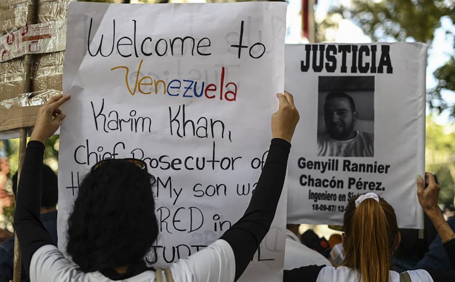 Deux manifestants brandissent des posters. Sur l'un, on peut lire en anglais : «bienvenue au Venezuela Karim Khan, procureur en chef de la CPI. Mon fils a été assassiné  lors d'une exécution extrajudiciaire.»
