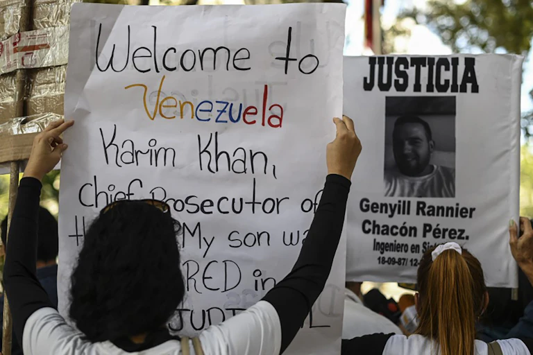 Deux manifestants brandissent des posters. Sur l'un, on peut lire en anglais : «bienvenue au Venezuela Karim Khan, procureur en chef de la CPI. Mon fils a été assassiné lors d'une exécution extrajudiciaire.»