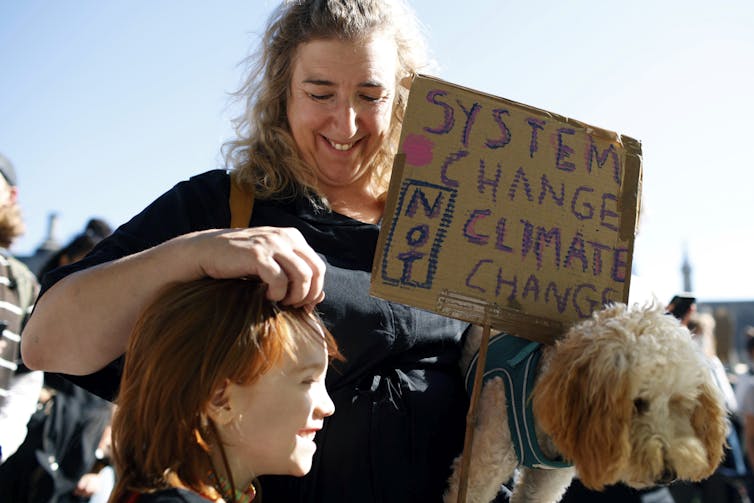 A woman rustles a child's hair as she holds a child-made sign that says 'systems change, not climate change.'
