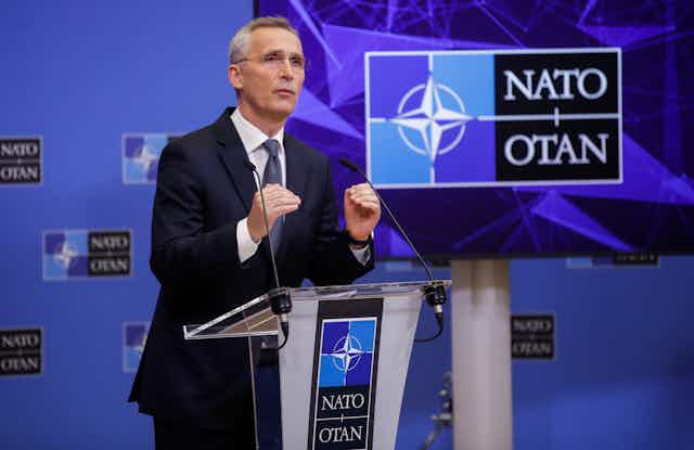 Man in suit speaks at podium in front of 'NATO' sign