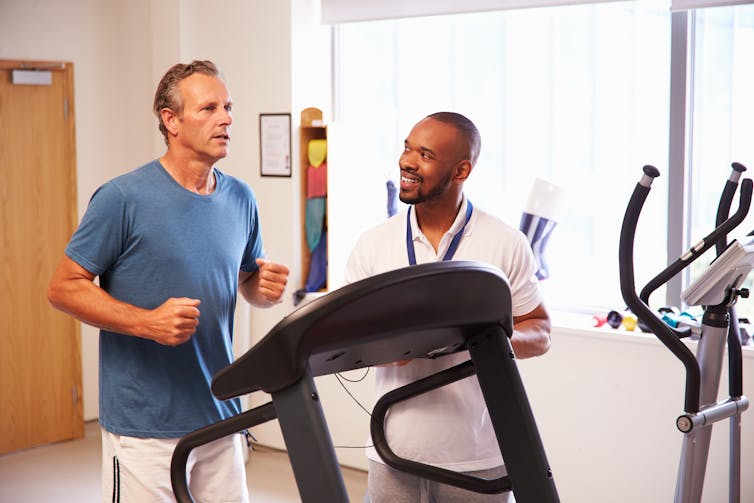 A man running on a treadmill while another man wearing a lanyard observes.