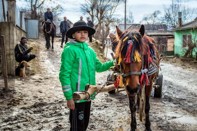 A boy in a black hat and green jacket stands beside a horse decorated with colourful tassles.