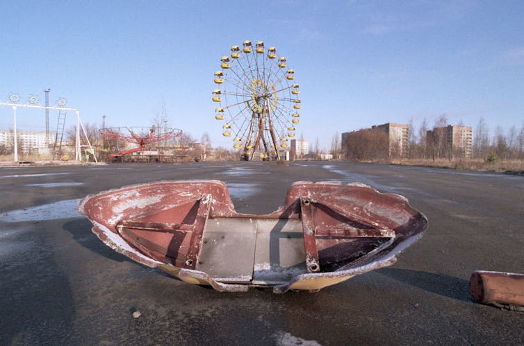 A derelict swing boat in an abandoned fun fair in front of a Ferris wheel.