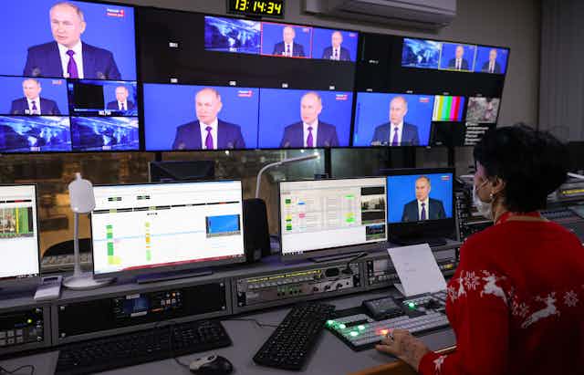 A woman sits facing a series of TV screens all showing Putin's face 