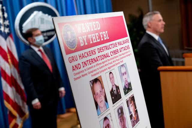 an FBI wanted poster mounted on a stand in the foreground with a man in a dark business suit and facemask standing in front of an American flag and another man in a dark business suit standing at a lectern in the background