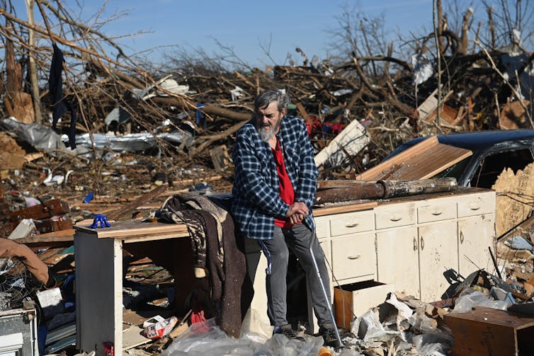 A man with a can sits on cabinets in what remains of his kitchen after a tornado. The roof and walls are gone.