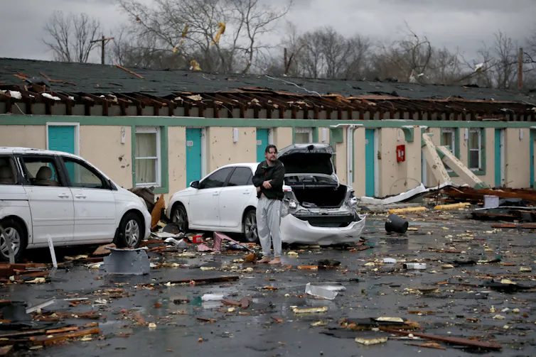 A man in sweatpants and no shoes appears distraught standing in the parking lot of a damaged motel.