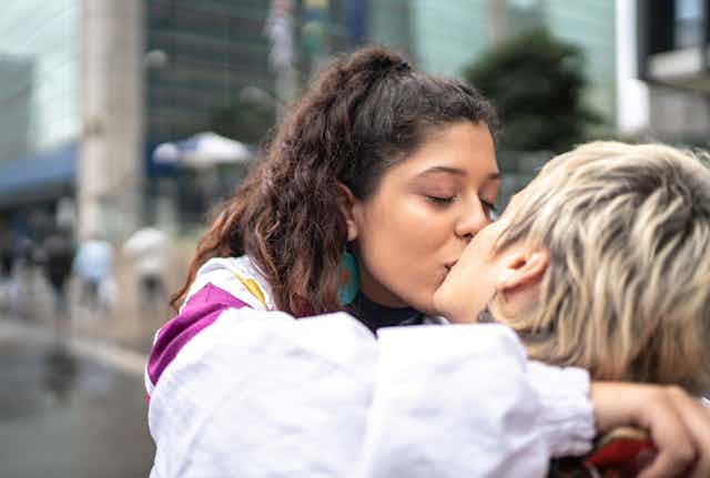A young woman with long dark hair kisses a person with short blonde hair on a busy street with people walking in background.