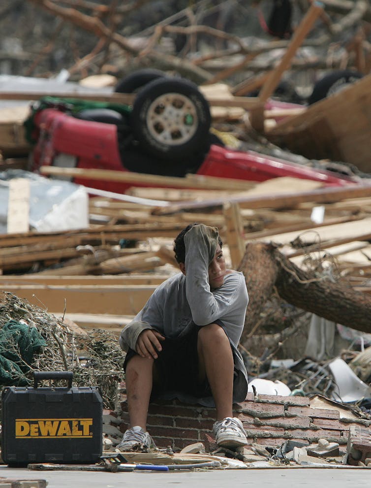 A boy with debris and an overturned car behind him.