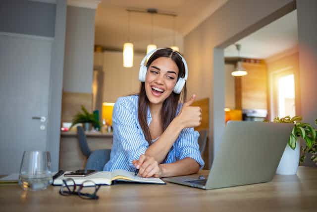 Mujer con auriculares ante un ordenador en una vivienda hace signo de acuerdo con el pulgar hacia arriba mientras sonríe.