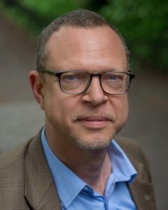 A portrait of a man in a blue shirt with a brown jacket, wearing glasses, his beard unshaven.