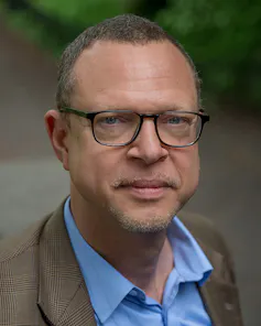 A portrait of a man in a blue shirt with a brown jacket, wearing glasses, his beard unshaven.