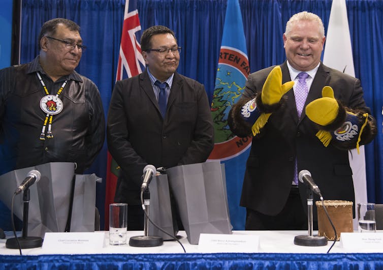 Chief Cornelius Wabasse, Chief Bruce Achneepineskum and Doug Ford stand behind a table in front of flags. Ford is wearing traditional leather gloves and clapping