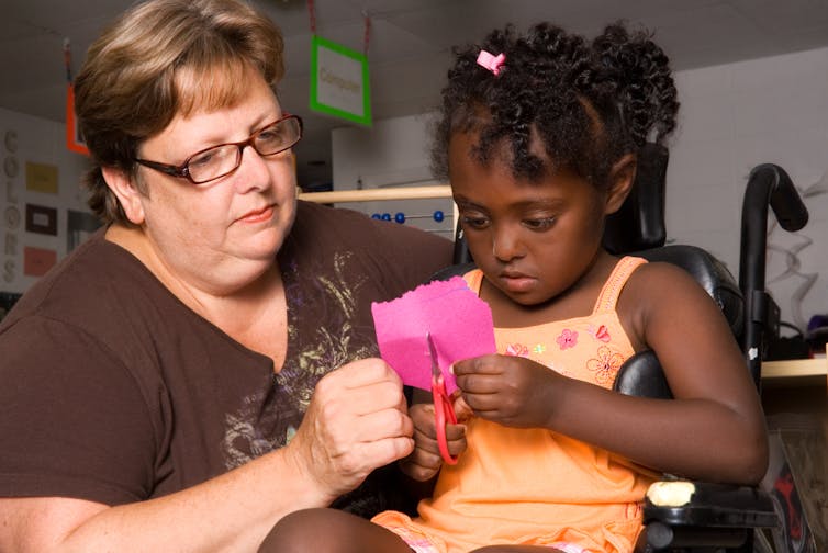 A teacher helps a child in a wheelchair cut a pink piece of paper with scissors.