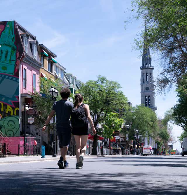 A couple walks down a treelined streed flanked by colourful buildings.