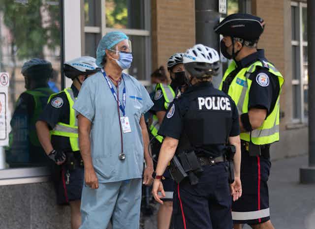 A health-care worker in blue scrubs standing outside with several uniformed police officers