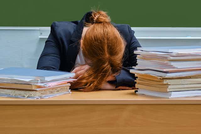 Exhausted teacher rests head on hands as she sits at her desk behind poles of work