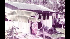 A woman in a colourful dress stands in front of a kitchen house with tropical trees around it.