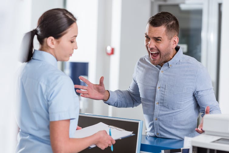 An angry-looking man in a blue shirt yelling at a health-care worker with a clipboard.
