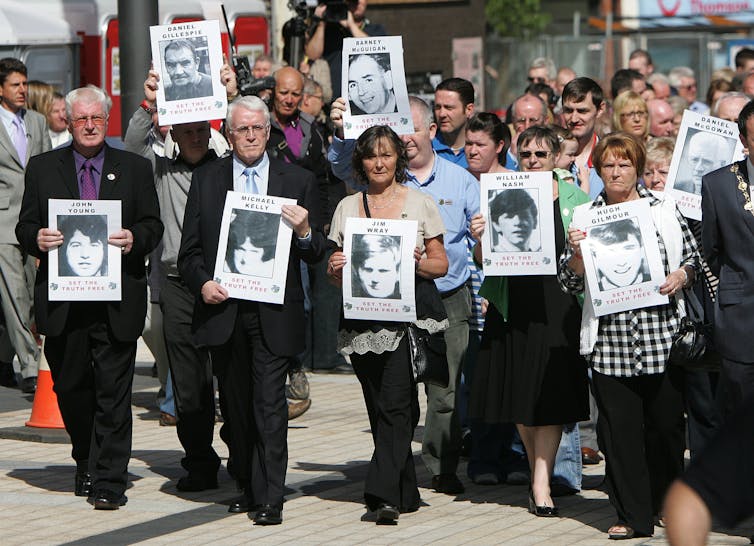 A large group of people marching are led by seven walking side by side and carrying black and white photos of their family members killed on Bloody Sunday.
