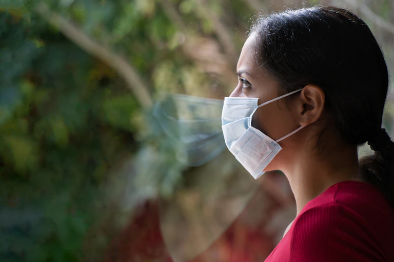 Una mujer con coleta y mascarilla de perfil justo a una ventana.