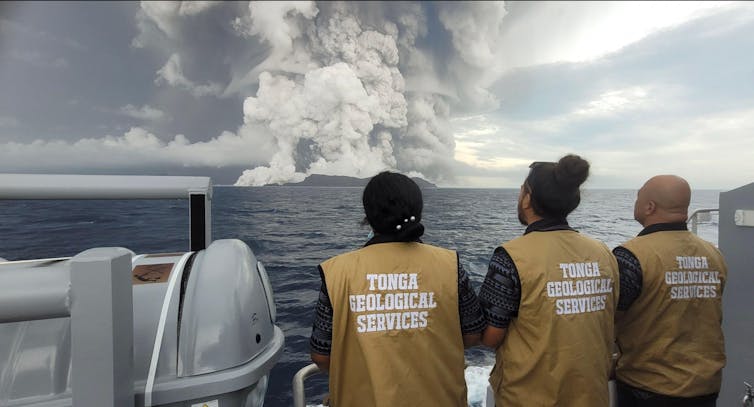 Three people with 'tonga geological services' jackets look at an eruption from a boat
