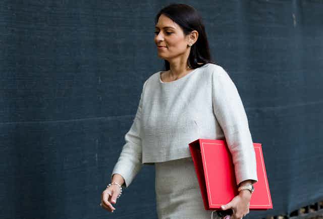 Home Secretary Priti Patel walks with a red folder under her left arm. She is wearing a matching grey top and skirt.