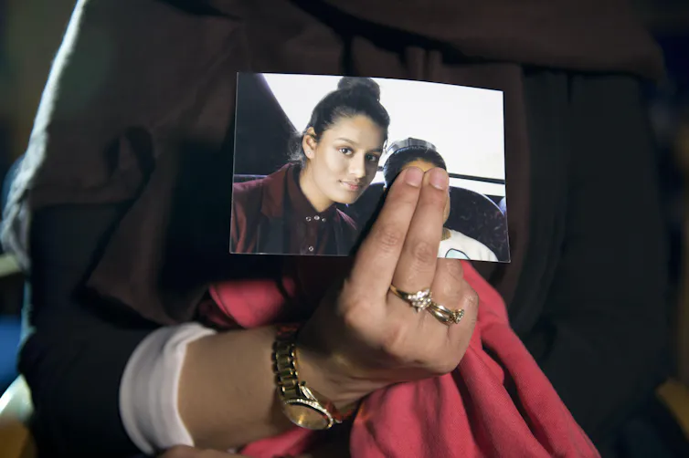 A close-up of a woman's hand holding a photograph of Shamima Begum. The woman is covering up the face of the other person in the photo with her fingers.