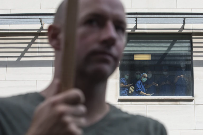 Health-care workers in masks peer out a hospital window at anti-vaccine demonstrators.