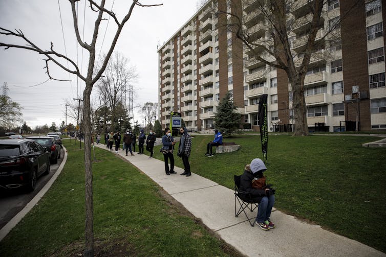 People line up along a sidewalk outside a brown apartment building. One man is sitting in a camping chair.